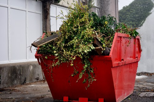 A large red industrial skip filled with garden waste including green branches, leafy foliage, and small woody stems, situated outdoors on a paved surface adjacent to a white fence with vertical panels, which appears to be part of a property boundary. The waste contains a mix of fresh green plant material and a few pieces of discarded wooden plank, with some leaves and twigs spilling over the edge of the skip. The background reveals a concrete wall with some climbing greenery and a partial view of neighboring buildings, indicating a residential or commercial area near Sydenham. The scene is set in natural daylight, providing clear visibility of the textures and colors of the waste and surroundings, contextualizing it within a waste collection or clearance operation performed by Office Clearance Sydenham for local rubbish removal services.