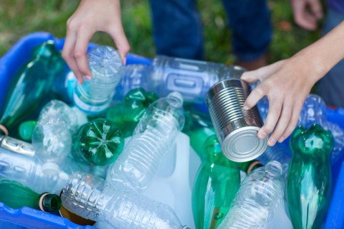 A close-up view of a person's hands, one holding a clear plastic water bottle and the other holding a cylindrical tin can, both positioned above a collection of discarded plastic bottles and containers. The plastic bottles are a mix of green and clear, some with labels still attached, and are placed inside a blue recycling bin. The background shows a grassy outdoor setting, indicating a recycling or rubbish collection activity, with natural daylight illuminating the scene. The image represents waste disposal related to plastic and beverage containers, consistent with rubbish removal services offered by companies like Office Clearance Sydenham in the Sydenham area.