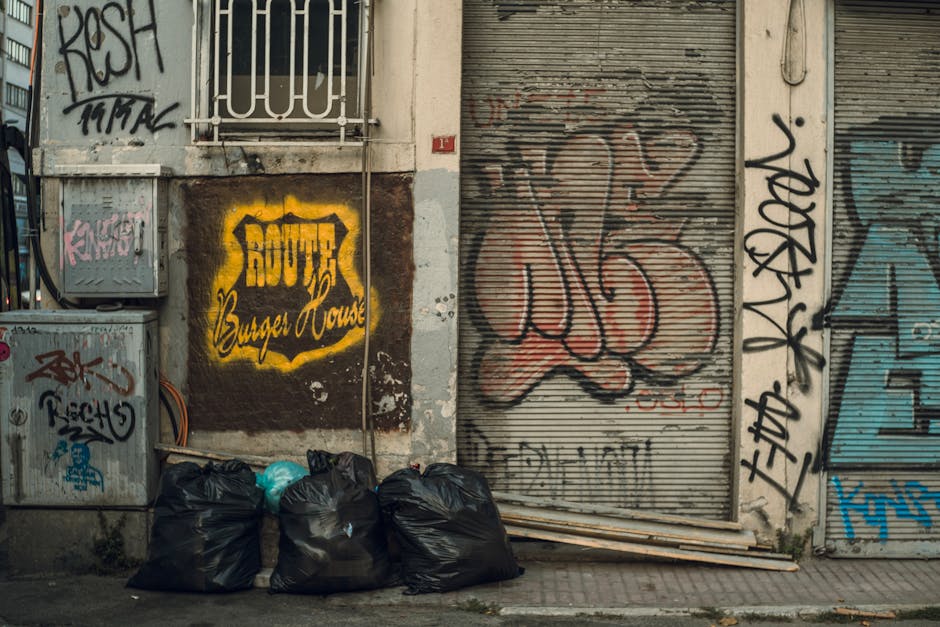 A large pile of mixed waste and rubbish bags is situated on a paved area adjacent to a street, with an open, overflowing grey recycling bin labeled for papers and cardboard positioned at the center. S