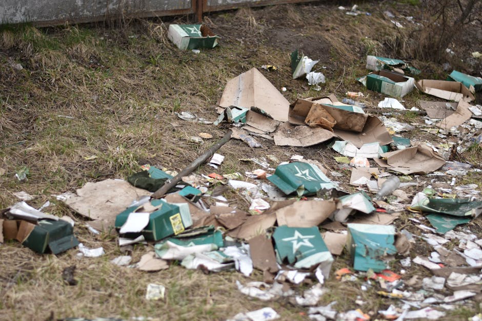 A pile of black rubbish bags and discarded materials, including pieces of cardboard and plastic, stacked against a weathered red metal door with graffiti tags in white paint located in an exterior all