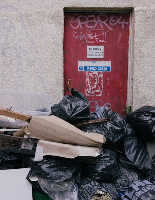 A photograph of a pile of assorted rubbish and waste materials situated on a concrete driveway outside a building. In the foreground, there are several large plastic bins filled with mixed items such